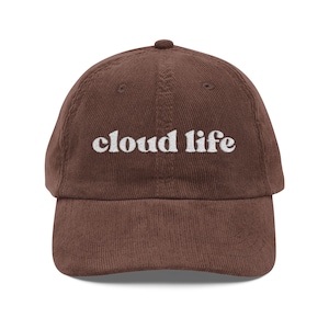 May include: A brown corduroy baseball cap with the embroidered text "cloud life" in white. The cap has a curved brim and a fabric-covered adjustable strap at the back. The hat is a casual accessory.