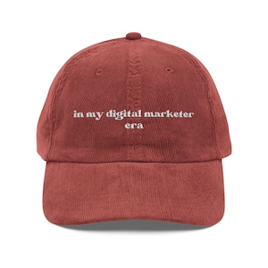 May include: A terracotta-coloured corduroy baseball cap with a curved peak. The cap has the white text "in my digital marketer era" embroidered on the front. The hat has a classic six-panel design with an adjustable strap.