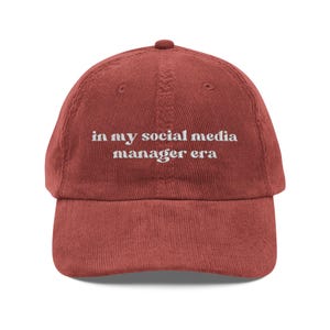 以下が含まれることがあります： レンガ色のコーデュロイ野球帽。白い文字で「in my social media manager era」と刺繍されています。帽子はカーブしたつばと調節可能なストラップが付いています。