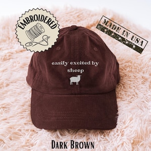 Puede incluir: Gorra de béisbol de pana marrón oscuro con la frase bordada "easily excited by sheep" y un gráfico de oveja. La gorra tiene una visera curva y un sello "Made in USA".
