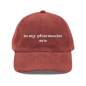 Puede incluir: Gorra de béisbol de pana rojo ladrillo con la frase "in my pharmacist era" bordada en blanco. La gorra tiene una visera curva y una correa ajustable.