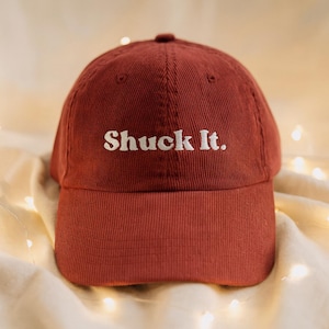 Peut inclure: Casquette de baseball en velours côtelé marron avec le texte "Shuck It." brodé en blanc. La casquette a une visière incurvée et une couronne arrondie. C'est un accessoire décontracté.