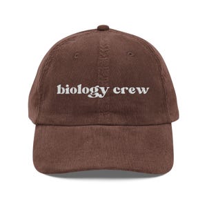 May include: A brown corduroy baseball cap with the embroidered text "biology crew" in white. The cap has a curved brim and a rounded top. The hat is a casual accessory.