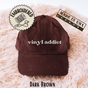 May include: Dark brown corduroy baseball cap with the text "vinyl addict" in white. The cap has a curved brim and a fabric strap closure. The image also includes an "Embroidered" patch and a "Made in USA" stamp.