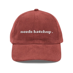 May include: A rust-colored corduroy baseball cap featuring the white embroidered text "needs ketchup." on the front panel. The hat has a structured crown, a curved brim, and a button at the top center, showcasing a textured fabric finish.