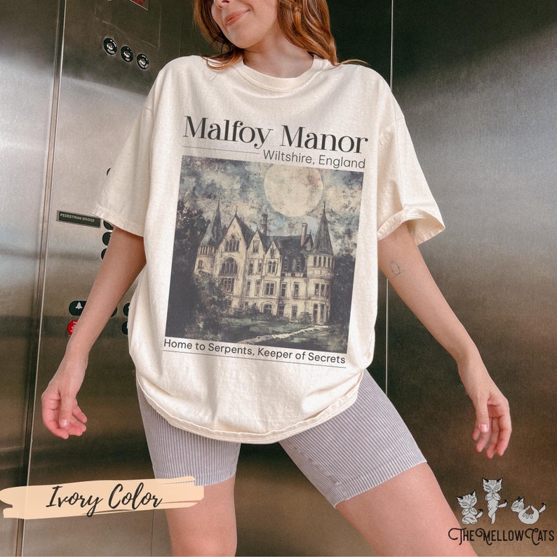 May include: Ivory coloured t-shirt featuring a graphic print of Malfoy Manor, a large, dark, gothic-style house with a full moon in the background. The text "Malfoy Manor, Wiltshire, England" and "Home to Serpents, Keeper of Secrets" is printed on the shirt.