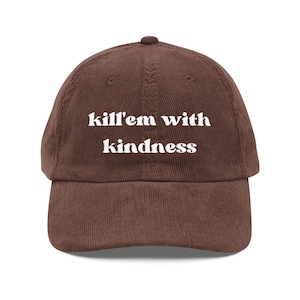 Könnte beinhalten: Braune Cord-Baseballkappe mit dem weißen Schriftzug "kill'em with kindness". Die Kappe hat einen gebogenen Schirm und einen Knopf oben. Die Kappe hat eine einfarbige braune Farbe.