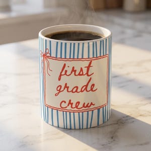 Pode incluir: Uma caneca de cerâmica branca com riscas verticais azuis e uma moldura retangular vermelha com um pequeno laço. O texto "first grade crew" está escrito em cursivo vermelho dentro da moldura e a caneca está cheia de café preto.