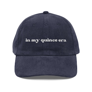 May include: A navy blue corduroy baseball cap featuring the embroidered white text "in my quince era" centred on the front panel. The hat has a structured crown, a curved brim, and a button at the top, showcasing a classic six-panel design.