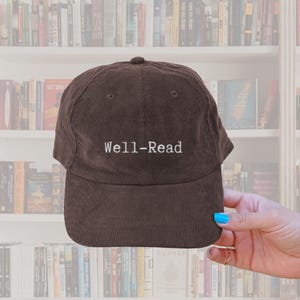 May include: A brown corduroy baseball cap with the embroidered text "Well-Read" in white. The cap has a curved peak and a rounded crown. The background shows a bookshelf filled with books.