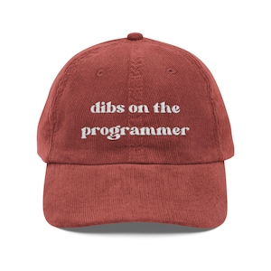 May include: A rust-coloured corduroy baseball cap featuring the embroidered white text "dibs on the programmer" centred on the front panel. The hat has a structured crown, a curved brim, and a button at the top, showcasing a textured fabric finish.