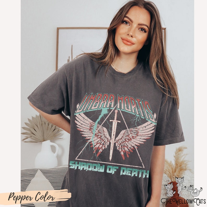 May include: A dark gray t-shirt with a vintage graphic print of a winged sword in a triangle with the text "Umbra Mortis Shadow of Death".