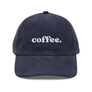 May include: A navy blue corduroy baseball cap featuring the word coffee. embroidered in white lowercase letters across the front centre. The hat has a structured crown, a curved brim, and a button at the top, showcasing a textured fabric finish.