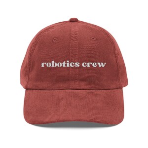 Peut inclure: Casquette de baseball en velours côtelé rouge brique avec le texte brodé "robotics crew" en blanc. La casquette a une visière incurvée et un bouton recouvert de tissu sur le dessus. Un cadeau pour les passionnés de robotique.