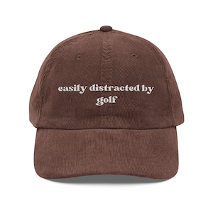 Puede incluir: Gorra de béisbol de pana marrón con la frase "easily distracted by golf" bordada en blanco. La gorra tiene una visera curva y una parte superior redondeada, ideal para actividades al aire libre.