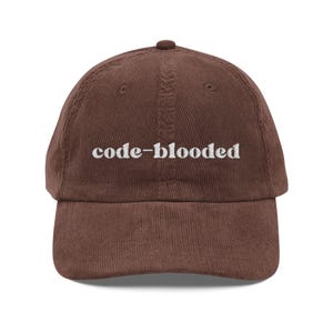 May include: A brown corduroy baseball cap with the text "code-blooded" embroidered in white. The cap has a curved brim and a button on top. The hat is a casual accessory.