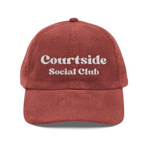 May include: A brick red corduroy baseball cap featuring the white embroidered text "Courtside Social Club" centered on the front panel. The hat has a structured crown, a curved brim, and a button at the top, showcasing a textured fabric finish.