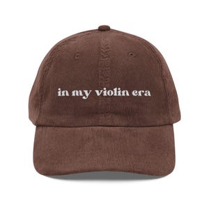 Könnte beinhalten: Braune Cord-Baseballkappe mit dem gestickten weißen Text "in my violin era". Die Kappe hat einen gebogenen Schirm und einen stoffbezogenen Knopf. Der Hut ist ein klassischer Stil, geeignet für Freizeitkleidung.