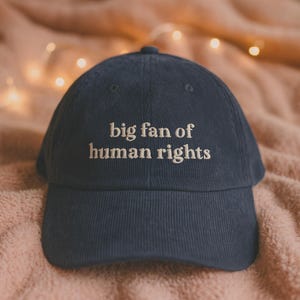 Könnte beinhalten: Marineblaue Cord-Baseballkappe mit dem gestickten Text "big fan of human rights" in Weiß. Die Kappe hat einen gebogenen Schirm und eine runde Krone. Der Hintergrund ist ein weicher, rosafarbener Stoff.