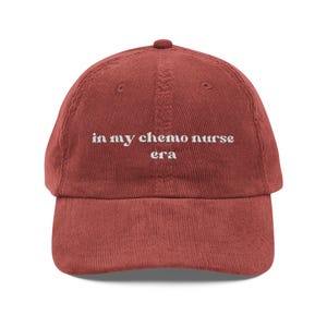 May include: A terracotta corduroy baseball cap featuring the white embroidered text "in my chemo nurse era". The cap has a curved peak and an adjustable strap at the back.