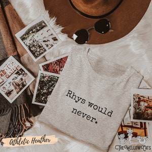 Puede incluir: Una camiseta gris con el texto "Rhys would never." impreso en ella. La camiseta est&aacute; sobre una superficie blanca y peluda con un sombrero marr&oacute;n, gafas de sol y varias fotos instant&aacute;neas.