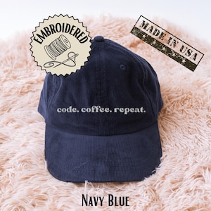 Puede incluir: Gorra de béisbol de pana azul marino con el texto bordado "code. coffee. repeat." y un sello "Made in USA". La gorra tiene una visera curva y un botón en la parte superior. La gorra está etiquetada como "Navy Blue".