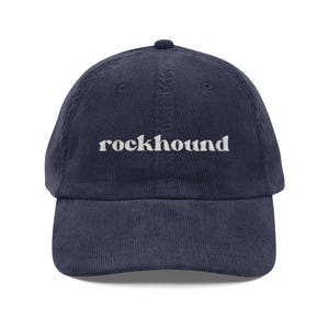 May include: A navy blue corduroy baseball cap featuring the word rockhound embroidered in white lowercase letters across the front center. The cap has a structured crown, a curved brim, and a button at the top of the dome with stitched eyelets.