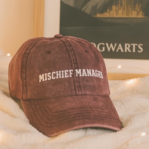 Peut inclure: Casquette de baseball marron avec les mots "MISCHIEF MANAGED" en lettres blanches. La casquette a une visière incurvée et un bouton sur le dessus. L'arrière-plan comprend une affiche avec le mot "GWARTS".