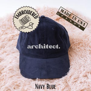 May include: Navy blue corduroy baseball cap with the word "architect" embroidered in white. The cap has a curved brim and a top button. The image also includes a "Made in USA" stamp and an embroidered design.