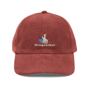 May include: A brick red corduroy baseball cap featuring an embroidered graphic of a white duck wearing a party hat, sitting on a red and white striped box with a blue star-patterned shape, and the text firequacker embroidered below it.