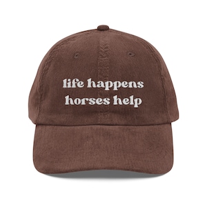 Könnte beinhalten: Braune Cord-Baseballkappe mit dem gestickten Text "life happens horses help" in Weiß. Die Kappe hat einen gebogenen Schirm und einen Knopf oben. Dieses Cap ist ein lässiges Accessoire.