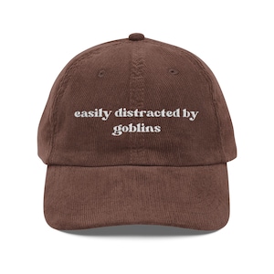 May include: A brown corduroy baseball cap with the text "easily distracted by goblins" embroidered in white. The cap has a curved brim and a fabric-covered adjustable strap.