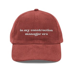 May include: A brick-red corduroy baseball cap with the embroidered text "in my construction manager era" in white. The cap has a curved brim and a button on top. The hat is a casual accessory.