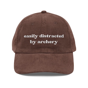 May include: A brown corduroy baseball cap with the embroidered text "easily distracted by archery" in white. The cap has a curved brim and a small button on top. This hat is a great gift for archers.