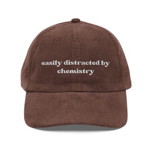 May include: A brown corduroy baseball cap with the embroidered text "easily distracted by chemistry" in white. The cap has a curved brim and a fabric-covered adjustable strap.