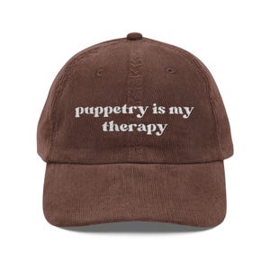 May include: A brown corduroy baseball cap with the phrase "puppetry is my therapy" embroidered in white. The cap has a curved brim and an adjustable strap at the back. The hat is a casual accessory.