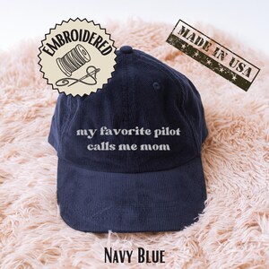 May include: Navy blue corduroy baseball cap with the embroidered text "my favorite pilot calls me mom." The cap has a curved brim and a stitched design. The words "Embroidered" and "Made in USA" are also visible.