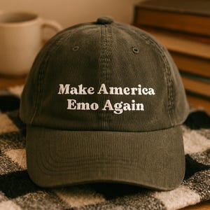 May include: Olive green corduroy baseball cap with the text "Make America Emo Again" embroidered in white. The cap has a curved brim and a fabric-covered button on top. The hat is sitting on a patterned surface.