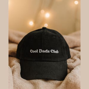 May include: A black corduroy baseball cap with the text "Cool Dads Club" embroidered in white. The cap is resting on a soft, textured surface, with blurred lights in the background.