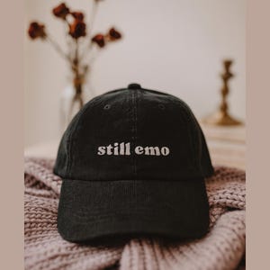 以下が含まれることがあります： 「still emo」の文字が白で刺繍された黒色のコーデュロイベースボールキャップ。ニットの表面に置かれています。背景にはドライフラワーの花瓶があります。