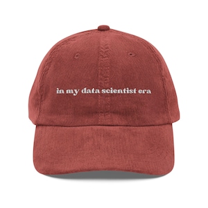 Könnte beinhalten: Ziegelrotes Cord-Baseballcap mit dem weißen Schriftzug "in my data scientist era". Die Kappe hat einen gebogenen Schirm und einen verstellbaren Riemen auf der Rückseite.