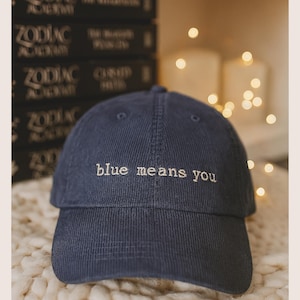 Puede incluir: Gorra de béisbol de pana azul marino con el texto bordado "blue means you" en blanco. La gorra tiene una visera curva y una corona estructurada. El fondo incluye libros e iluminación suave.