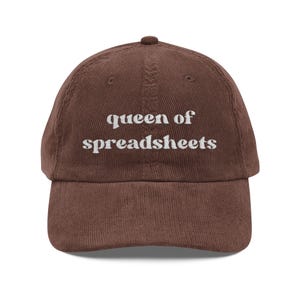 May include: A brown corduroy baseball cap with the embroidered text "queen of spreadsheets" in white. The cap has a curved brim and a button on top. The hat is a casual accessory.