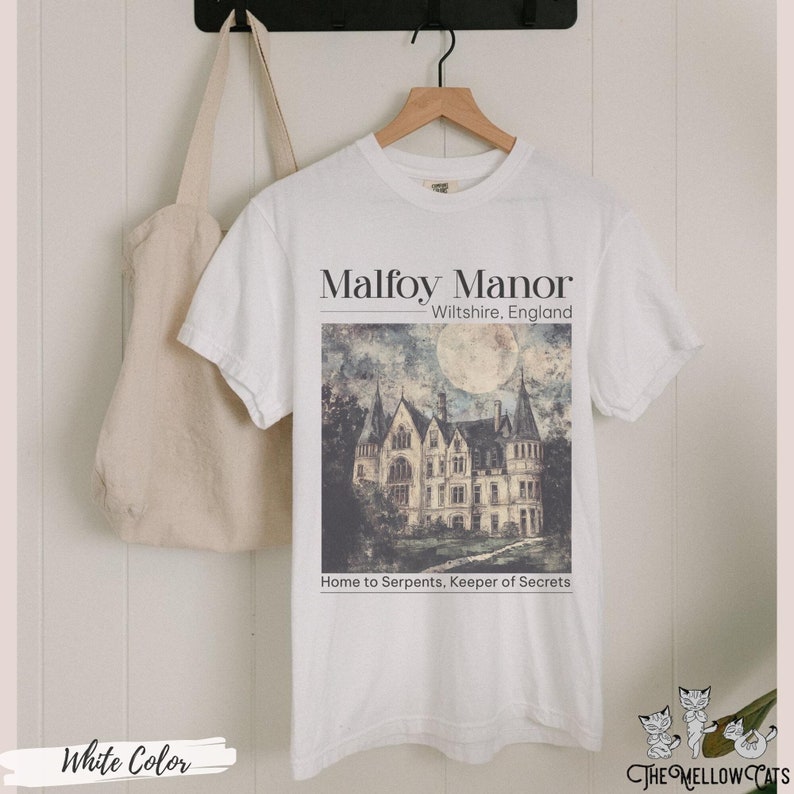 May include: White t-shirt featuring a graphic of a large, grey stone manor house with a full moon in the sky. The text "Malfoy Manor" is above the image and "Wiltshire, England" is below the image. The text "Home to Serpents, Keeper of Secrets" is below the manor house.