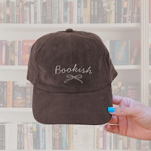 May include: A brown corduroy baseball cap featuring the embroidered word "Bookish" in white script above a bow. The cap has a curved peak and a button on top. The background is a blurred image of a bookshelf.