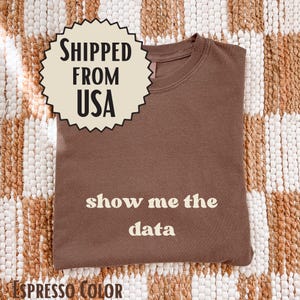 Könnte beinhalten: Ein einfarbiges, kurzärmeliges T-Shirt in Espressobraun mit dem Text "show me the data", der in einer cremefarbenen, abgerundeten Serifenschrift mittig auf der Brust aufgedruckt ist. Das Kleidungsstück hat einen klassischen Rundhalsausschnitt.