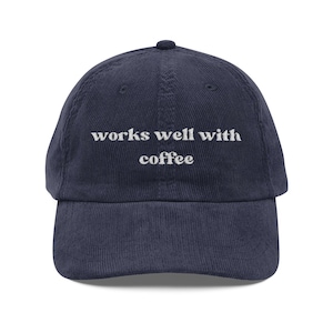 May include: A navy blue corduroy baseball cap featuring the white embroidered text "works well with coffee" centred on the front panel. The hat has a structured crown, a curved brim, and a button at the top, showcasing a classic six-panel design.