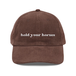 May include: A brown corduroy baseball cap with the phrase "hold your horses" embroidered in white across the front. The cap has a curved peak and a rounded top.