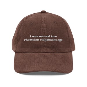 May include: A brown corduroy baseball cap with the text "i was normal two rhodesian ridgebacks ago" embroidered in white. The cap has a curved brim and a button on top.