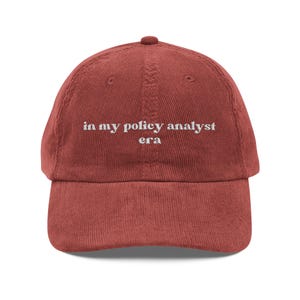 May include: A brick-red corduroy baseball cap with a curved brim. The cap features the embroidered text "in my policy analyst era" in white, centered on the front. The hat has a classic six-panel design with a top button.